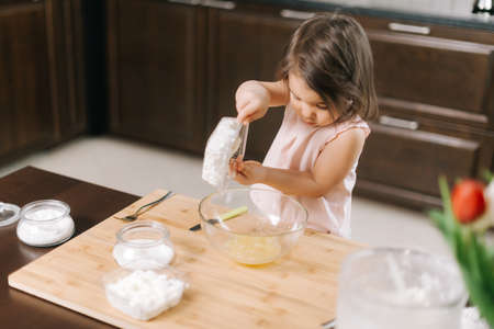 Attractive little girl puts the cottage cheese in a bowl of whipped eggs to make cheesecakes.