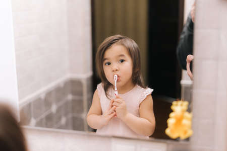 Adorable beautiful little girl is brushing her teeth with tooth brush in front of the bathroom...