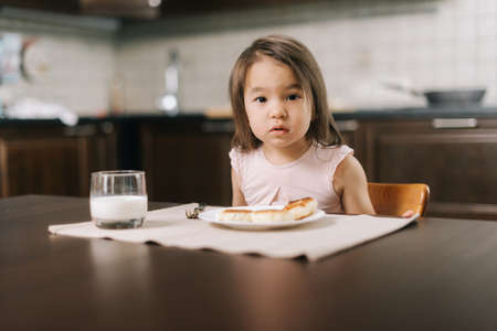 Beautiful little two-year-old girl is waiting for the meal to begin, looking at the camera.. Cheesecakes on the table in front of the child and a glass of milk for breakfast.の写真素材