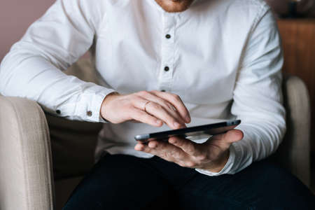 Close-up of a businessman in white stylish shirt working on digital tablet, sitting in chair in...