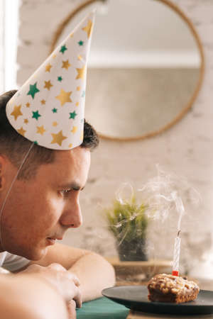 Thoughtful sad young man sitting in front of little cake with lighted candle and blowing it, close-up of face.の写真素材