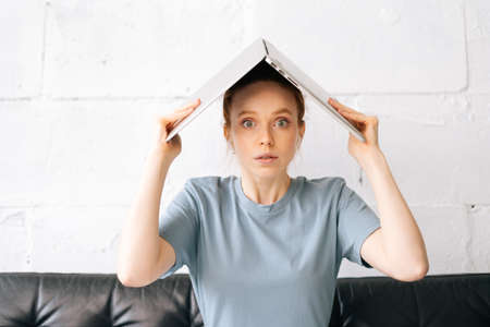Shocking redhead young woman sitting on couch and put the book on head in cozy room at home, looking at camera. Unhappy student with book on the head sitting at sofaの写真素材