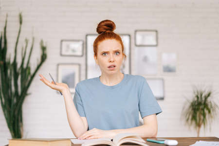 Surprised disappointed redhead young woman student is sitting at a table with books and a workbook, looking at camera. Girl student preparing for college exams.の写真素材