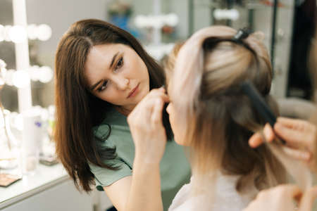 Close-up of preparation process of gorgeous young woman, hairdresser is twisting long fair hair, female make-up artist working in dressing room. Concept of backstage work.の写真素材