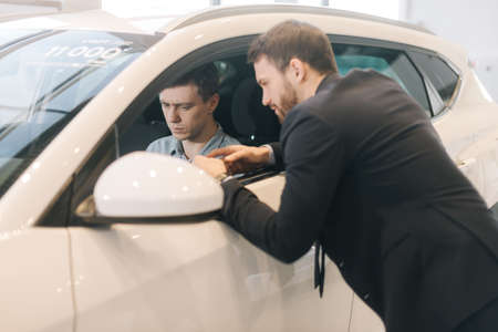 Handsome young man testing of new car in auto dealership. Professional car salesman wearing business suit is telling about new car model. Concept of choosing and buying new car at showroom.の写真素材