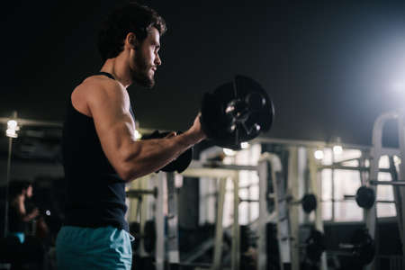 Handsome bearded young man with muscular wiry body wearing sportswear working out with barbell during sport workout training in modern dark gym. Concept of healthy lifestyle.の写真素材