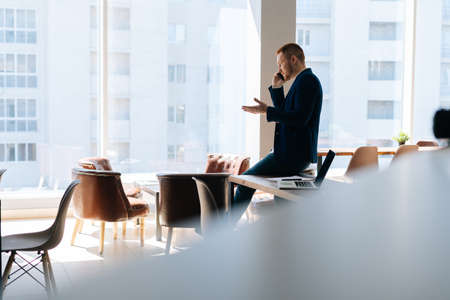 Serious young businessman wearing fashion suit is talking on mobile phone in modern office room near wooden desk on background of large window. Concept of office working.の写真素材