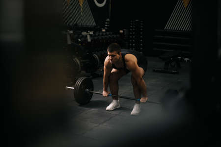 Muscular fit man with perfect beautiful body wearing sportswear lifting heavy barbell from floor during sport workout training in modern dark gym. Concept of healthy lifestyle.の写真素材