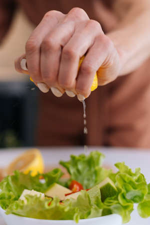 Close-up of womens hands squeezing juice from fresh yellow lemon into salad bowl with sliced...