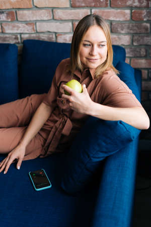 Portrait of gorgeous blonde woman wearing fashion clothing, holding fresh green apple, while lying...