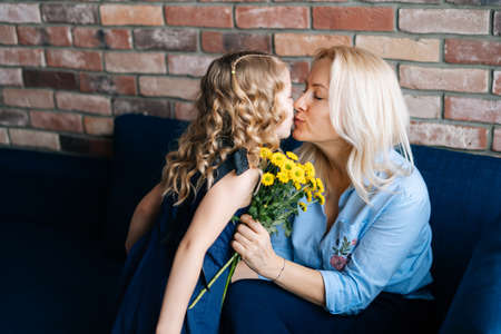 Daughter and mother are kissing in their home, girl gives mother a bouquet of flowers in cozy living room. Concept of Happy Mothers Day.の写真素材