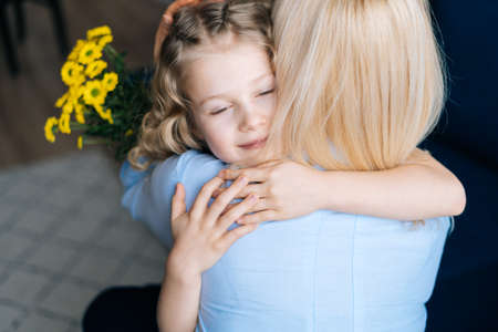 Cute mother and her adorable little daughter are hugging at the home in cozy living room. Loving daughter gives bouquet flowers to her mother.Concept of Happy Mothers Day.の写真素材