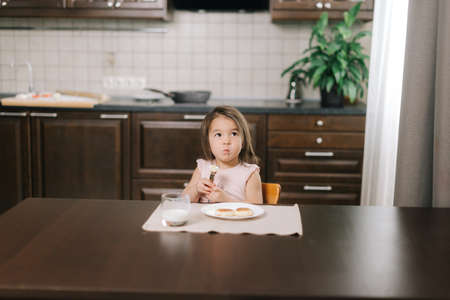 Happy beautiful little two-year-old girl is eating tasty cheesecake with fork for dinner at the table in kitchen room. Glass of milk on the table.の写真素材