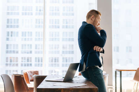 Side view of thoughtful young business man wearing fashion suit clothing is standing in modern office room near wooden desk on background of large window. Concept of office working.の写真素材