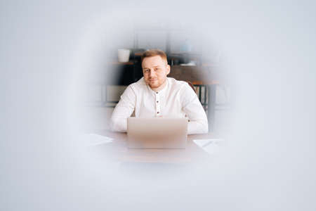 Remote view of busy young business man wearing fashion casual clothing is working on laptop computer at the table in modern office room ,looking at the camera.. Concept of office working.の写真素材