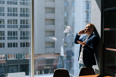 Happy young businessman wearing fashion suit is talking on mobile phone in modern office room near large window. Concept of office working.の写真素材