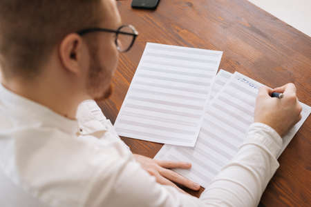 Side view of talented elegance young composer wearing stylish eyeglasses writing musical notes with pen on paper sheet music sitting at the wooden desk in modern room in home or office.の写真素材