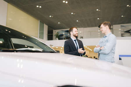 Happy smiling customer and friendly salesman wearing business suit are discussing new car in auto dealership. Concept of choosing and buying new car at showroom.の写真素材