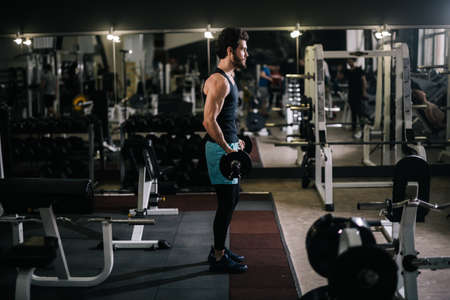 Sportive bearded young man with muscular wiry body wearing sportswear holding barbell during sport...