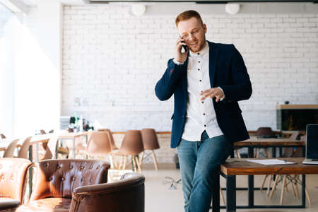 Happy young businessman wearing fashion suit is talking on mobile phone in modern office room near wooden desk Concept of office working.の写真素材