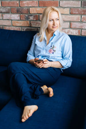 Relaxed young blonde woman using cell phone while sitting on soft dark blue sofa in living room at home office. Attractive female housewife sitting on couch and looking at away. Casual style portraitの写真素材