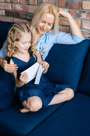 Young blonde mother and her curly little girl look through the photos while sitting on the soft sofa...