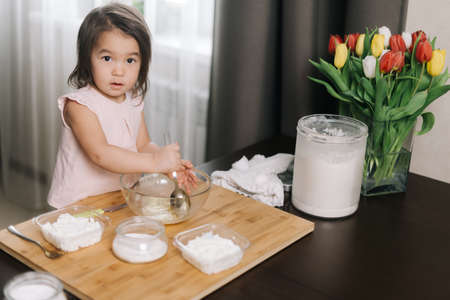 Little two-year-old girl wearing white dress is using stainless spoon to mix the egg for making bakery at kitchen, looking at the camera.の写真素材