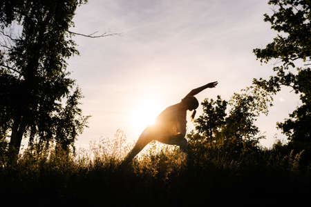 Silhouette of unrecognizable woman practicing yoga asana pose of warrior in evening at sunset. Girl yoga instructor performing Virabhadrasana posture outside. Concept of practicing yoga alone outdoor.の写真素材