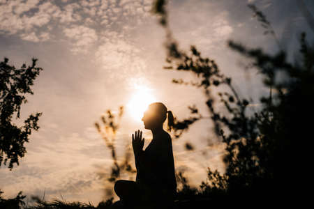 Silhouette of young woman meditating in lotus position with closed eyes, from below. Relaxed lady is performing namaste yoga pose background of sun outdoor on park outside city evening during sunset.の写真素材