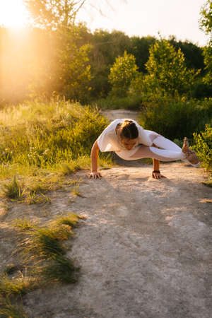 Young woman with stretching body practicing yoga and performing Eight-Angle Pose outside in park evening on background of sunlight. Female doing advanced yoga exercises outdoors at green grass.の写真素材