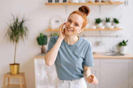 Portrait of happy carefree redhead young woman listening music in wireless headphones while sitting...