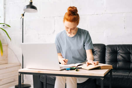 Portrait of redhead young woman student noting into the workbook important information from the book...
