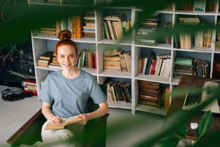 Happy redhead young woman student is reading book enjoys of rest at home office, looking at camera. Cute lady enjoying books at library room on background of bookshelves.の写真素材