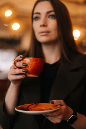 Close up of face of young woman holding cup of tasty hot coffee in hand while standing in restaurant. Caucasian lady posing with glass of beverage in cafe with modern dark interior.の写真素材