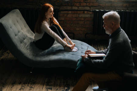 Professional gray-haired man psychologist consulting patient during psychological consultation in cozy dark office room. Bearded mature psychotherapist taking notes and giving advices to patient.の写真素材