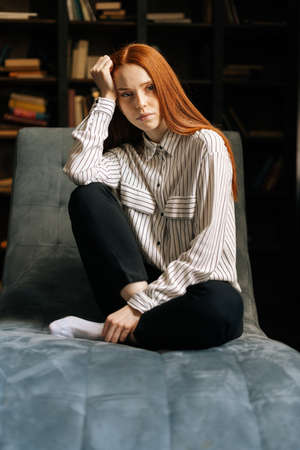 Sad attractive red-haired young woman sitting in chair and looking at the camera on background of...
