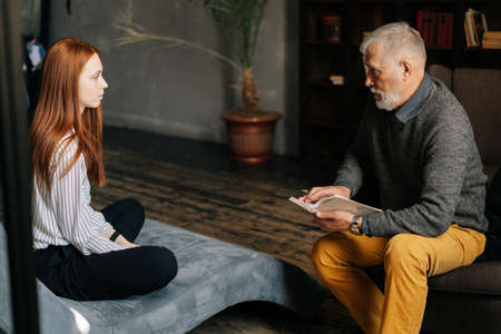 Side view of red-haired young woman patient speaking with mature man psychologist. Skilled psychotherapist asking questions and writing on clipboard. Concept of psychological treatment.の写真素材