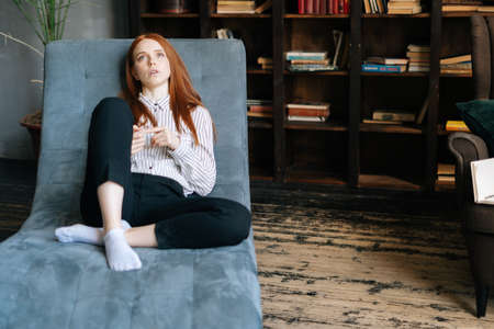 Portrait of sad red-haired young woman talking about problems lying in armchair in psychological unloading room. Depressed lady at psychologist appointment. Concept of psychological treatment.の写真素材
