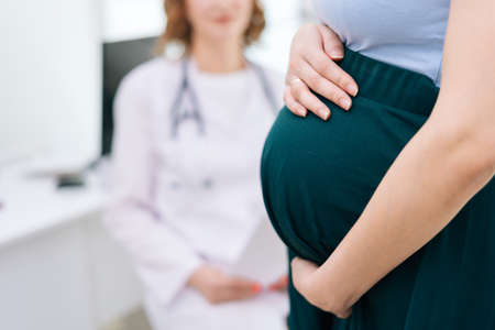Close-up of pregnant woman holding hands on belly while sitting in tight medicine office. Female doctor standing on background. Concept of happy and healthy childbearing.の写真素材