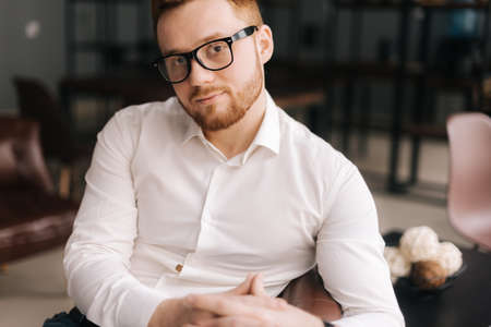 Creative young business man wearing stylish glasses in trendy clothing looking at camera while sitting indoors on chair in modern office. Portrait of bearded handsome gentleman wearing white shirt.の写真素材