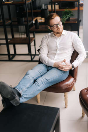 Relaxed young business man wearing stylish glasses in trendy clothing looking away while sitting indoors on the chair in modern office. Portrait of bearded handsome gentleman wearing white shirt.の写真素材