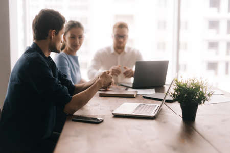Young business man tells his partners about the project in modern office meeting against large panoramic window. Young man and woman listen to the speaker. Concept of office life.の写真素材