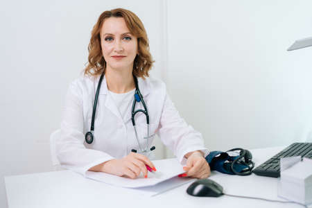Attractive blonde young woman doctor sitting in white office and looking at the camera. Portrait of therapist wearing white coat with stethoscope posing on white office room.の写真素材