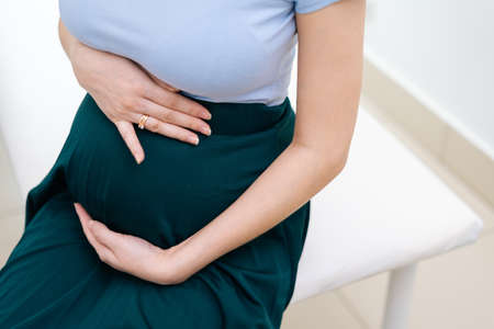 Close-up of pregnant woman holding hands on belly while sitting in tight medicine office. Concept of...