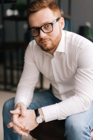 Serious young business man wearing stylish glasses in trendy clothing looking at camera while sitting indoors on chair in modern office. Portrait of bearded handsome gentleman wearing white shirt.の写真素材