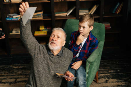 Caucasian cheerful bearded gray-haired grandfather with his grandson enjoy memories watching family photo album sitting on armchair and floor at home in room with an authentic aristocratic interior.の写真素材