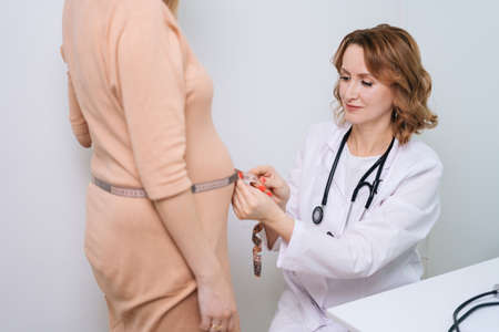 Smiling woman doctor wearing white coat is measuring waist size of pregnant young female using measuring tape in white office room. Concept of happy and healthy childbearing.の写真素材