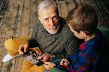 Top view of handsome bearded gray-haired grandfather with his grandson having fun looking at an old photo album, enjoying memories watching family photo album at home in living roomの写真素材