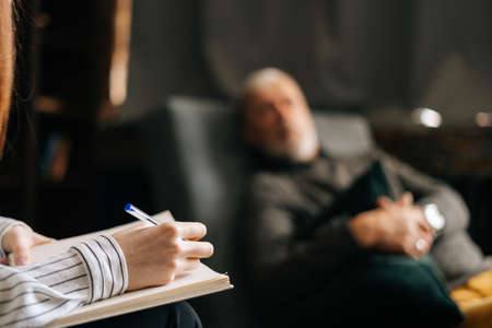 Close up view of woman psychologist taking notes in notebook, mature man patient answering questions in office passing psychological test. . Concept of psychological treatment.の写真素材