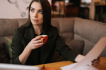 Focused young business woman studying paper documents in cafe sitting at table with laptop. Lady enjoys delicious coffee while working. Student girl sitting in cafe with cup of coffee.の写真素材